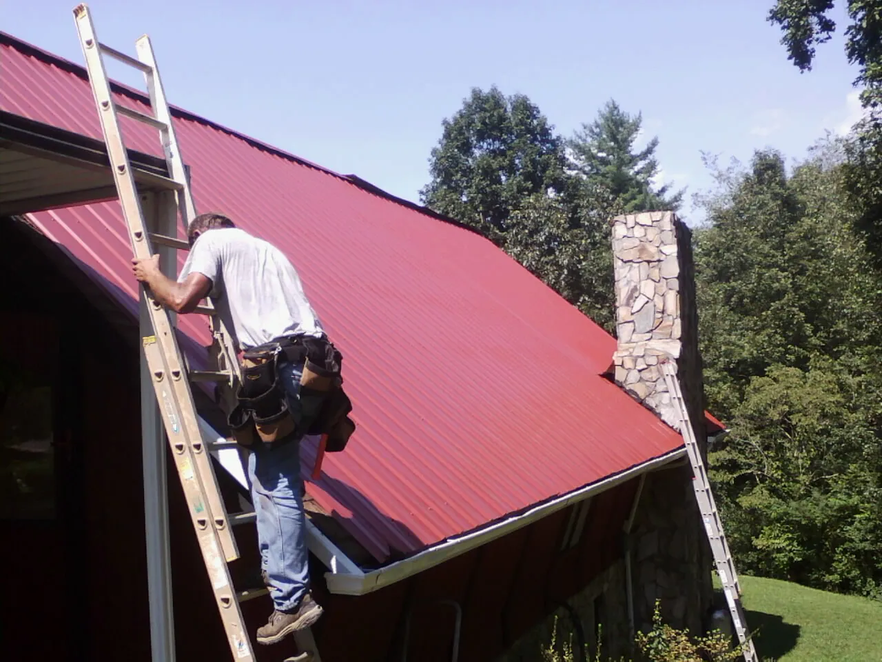 Roofer on a ladder beside a red metal roof and stone chimney — Soffit & Fascia Repair by Roofing Brothers in Genoa City